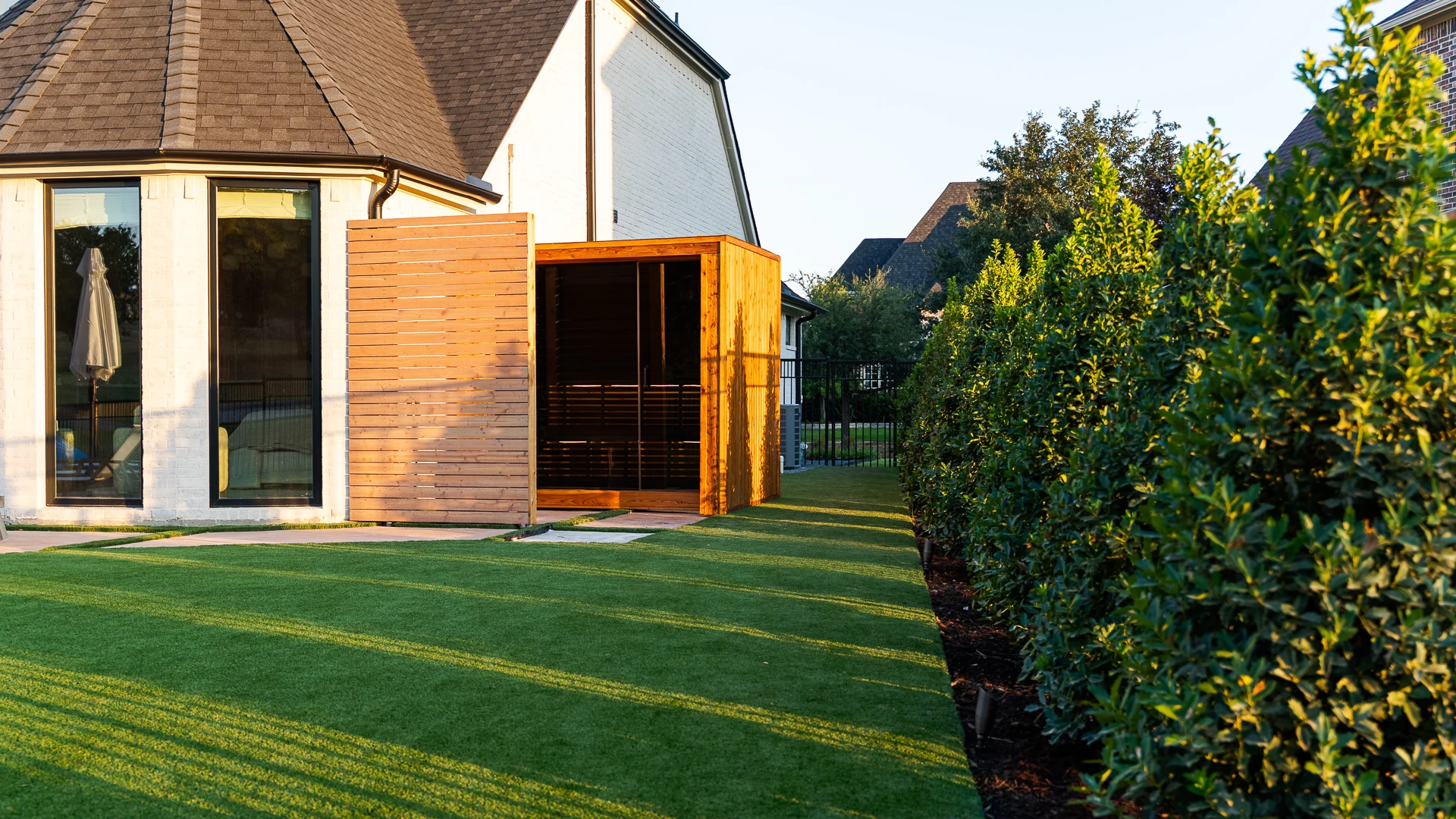 Cedar cube sauna with slat privacy screen on artificial turf at golden hour