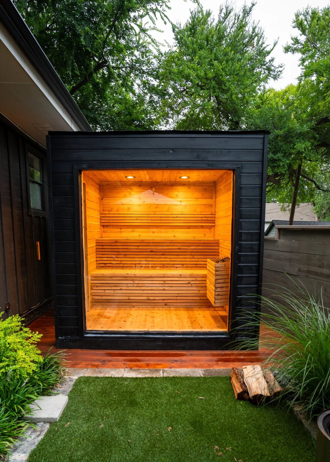 Black outdoor sauna cube with glowing cedar interior seen through full-height window